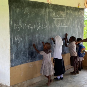 4 Kinder stehen an einer Tafel und schreiben. Hinter ihnen steht die Lehrerin und erklärt etwas. Auf der Tafel stehen verschiedene Wort auf Kisuaheli geschrieben.