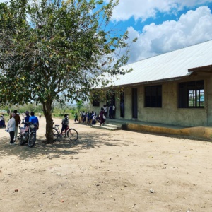 Das Gebäude ist die Komkomba Schule, davor steht ein Baum, in seinem Schatten stehen einige Schulkinder